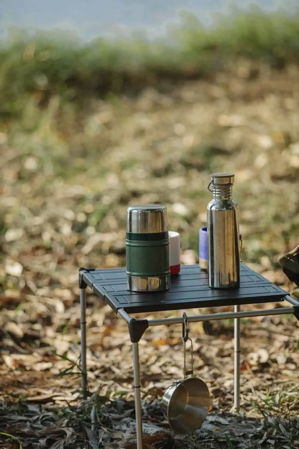 A folding camp table holds a thermos, a steel water bottle, and a cup, with a pan hanging from a hook underneath.
