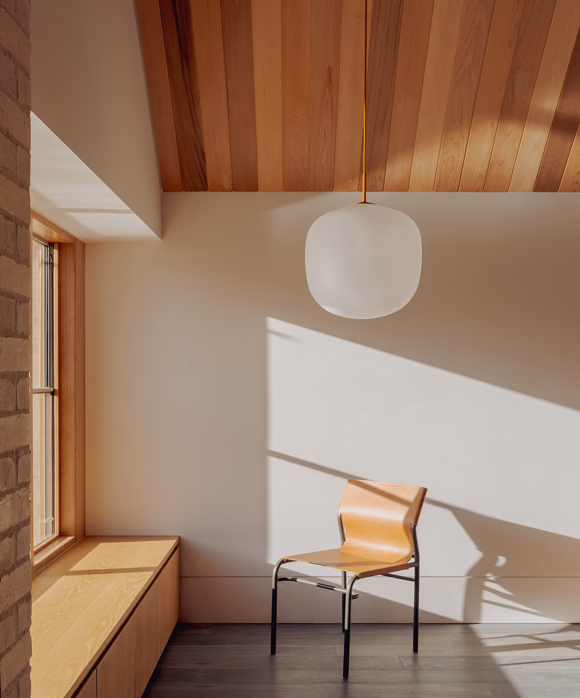 Minimalist room with wooden ceiling, window bench, and a modern chair under a pendant light.