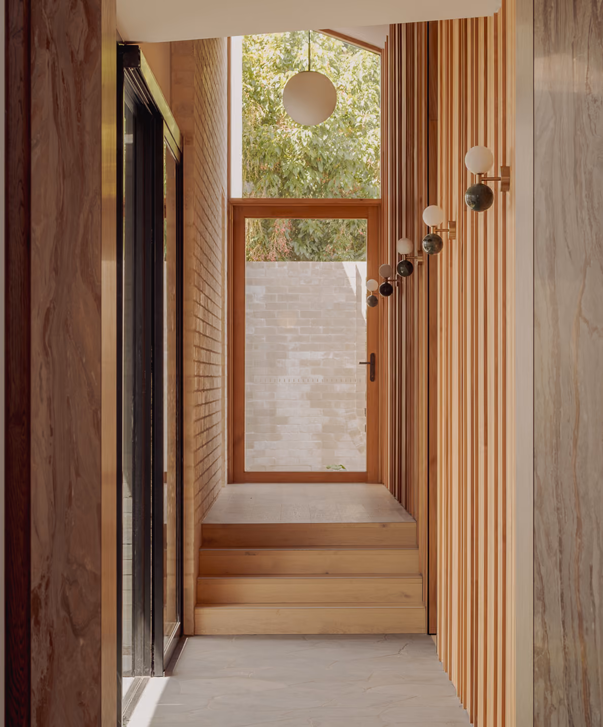 Modern hallway with wooden steps, vertical wood paneling, round wall lights, and a glass door with tree view.