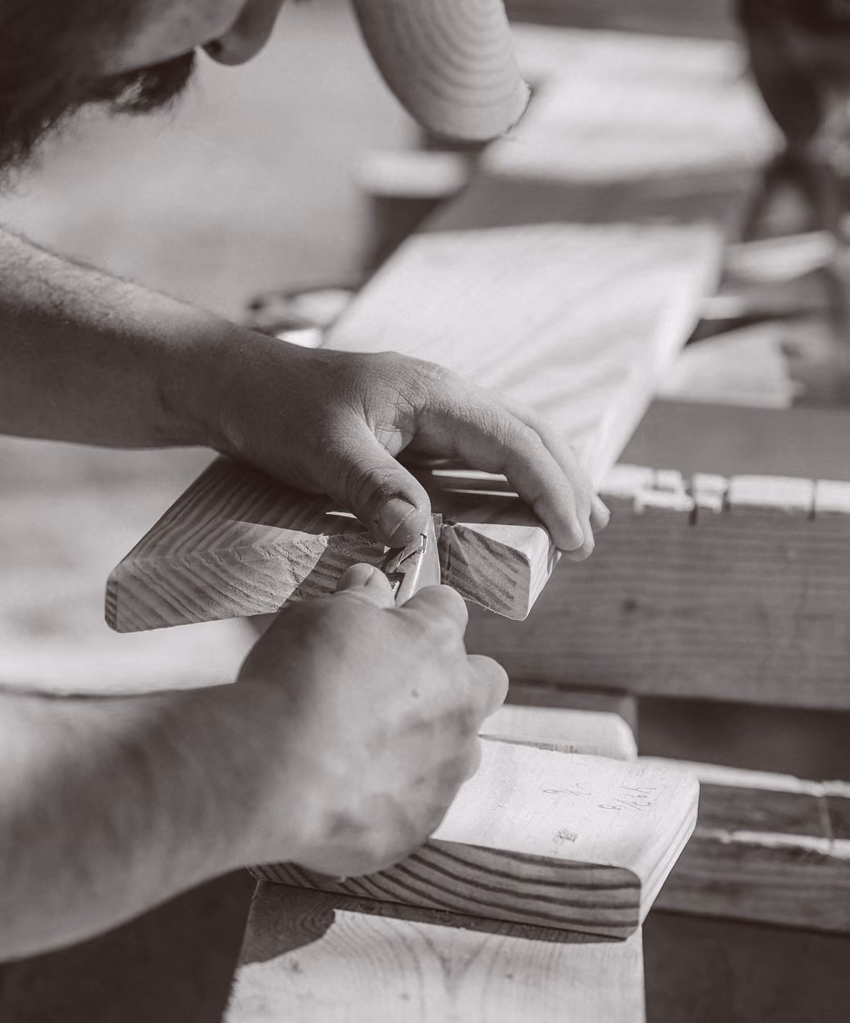 Person carving or shaving the edge of a wooden plank using a hand tool on a workbench.