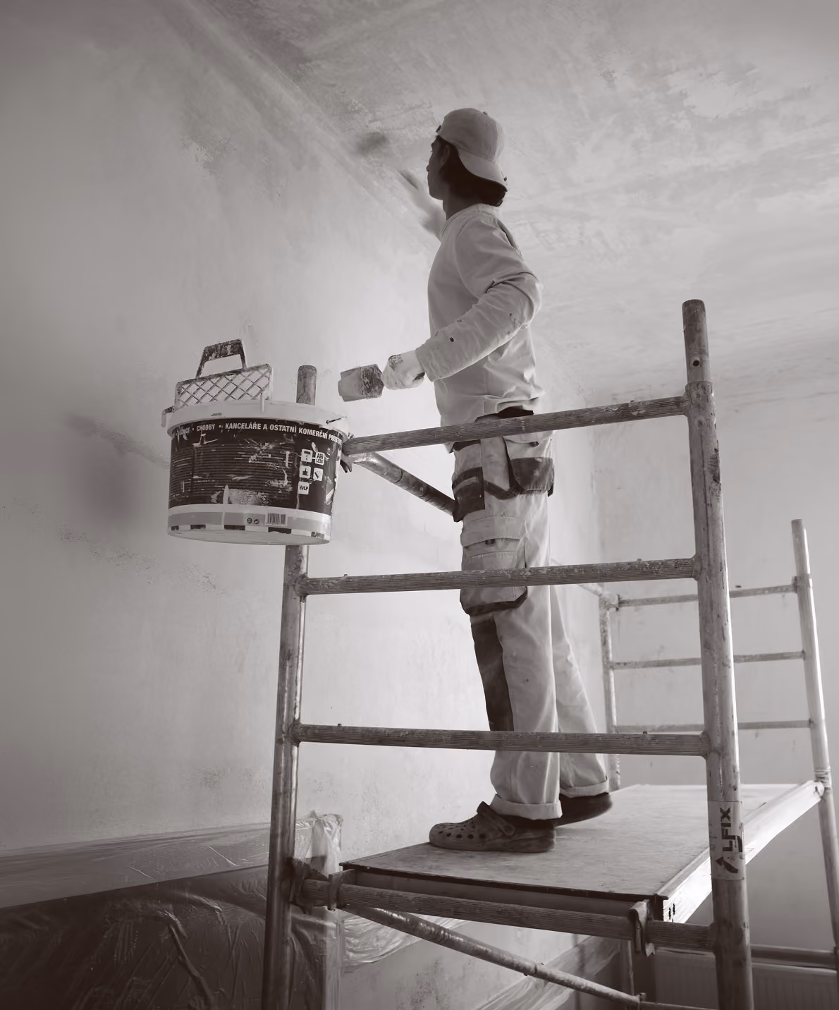 Person standing on scaffold painting a ceiling with a brush and holding a paint bucket.