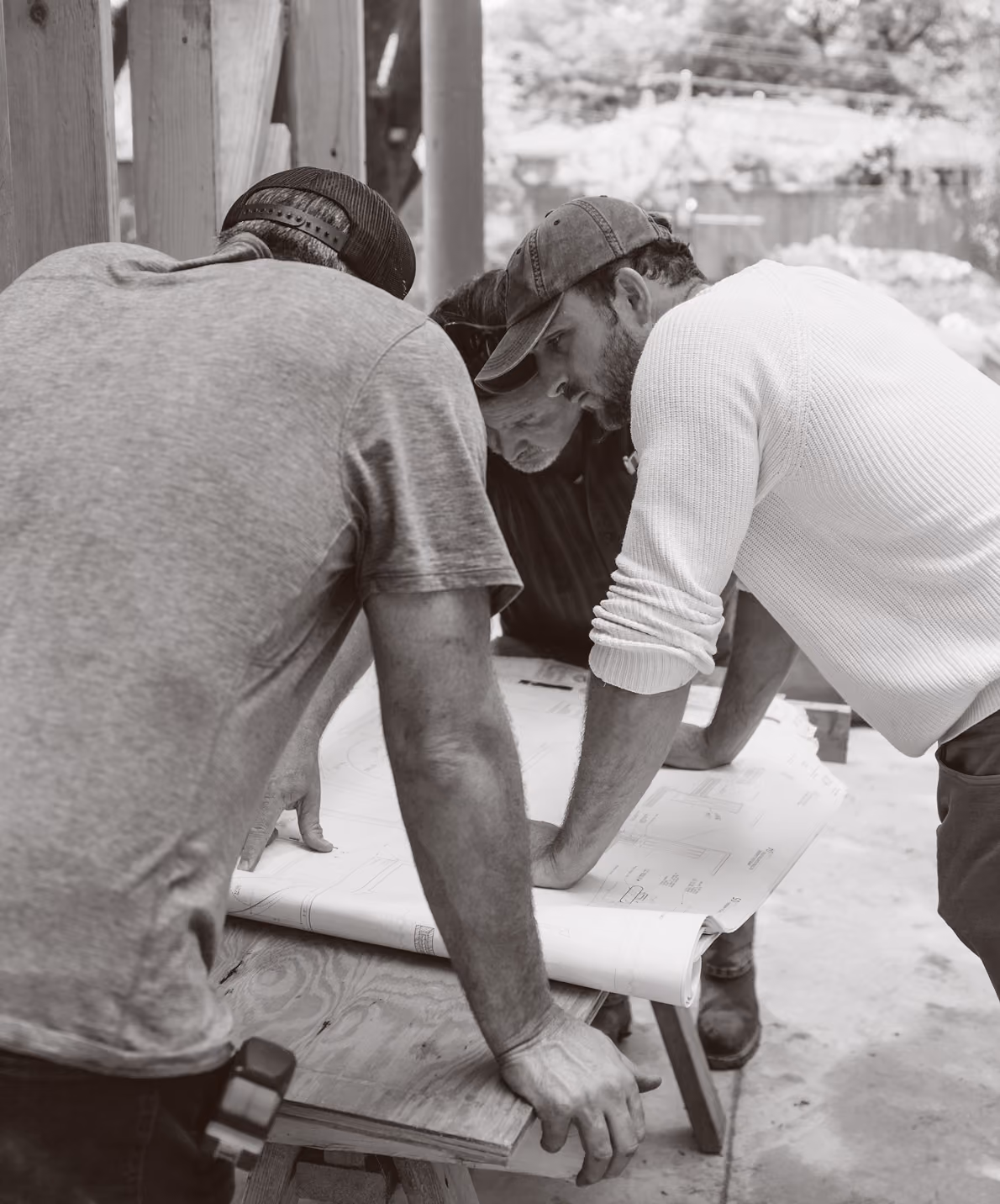 Three men in caps lean over a table reviewing a large blueprint at an outdoor worksite.