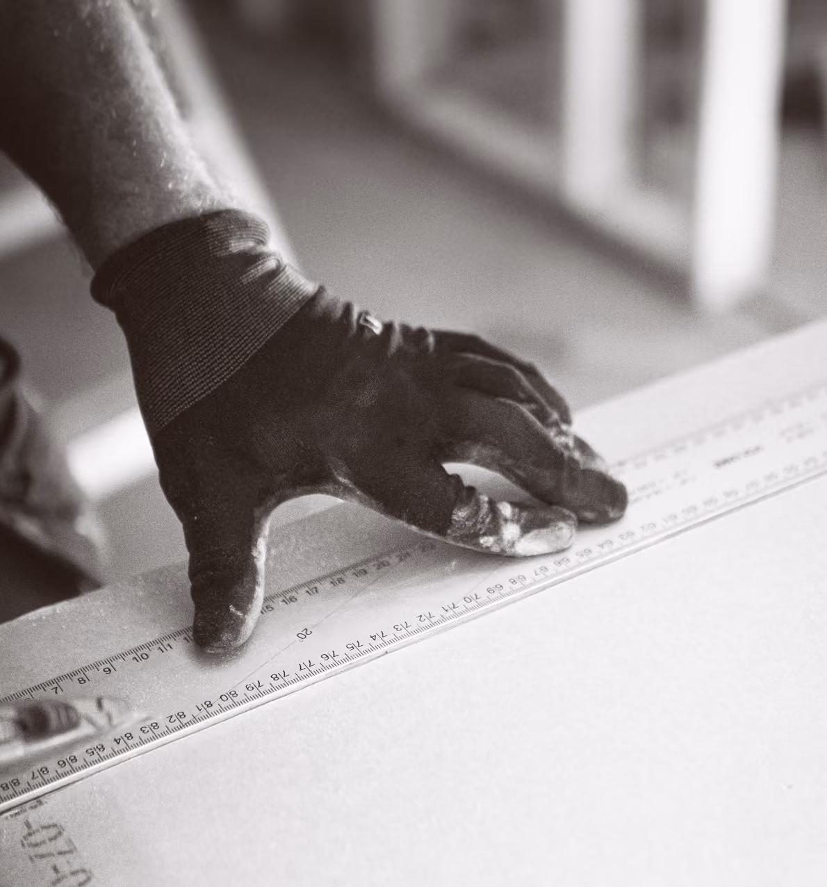Hand wearing a glove holding a ruler on a flat surface, likely measuring or marking it.