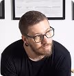 Man with short hair, beard, glasses, and black shirt looking slightly down and to the side in front of framed documents.