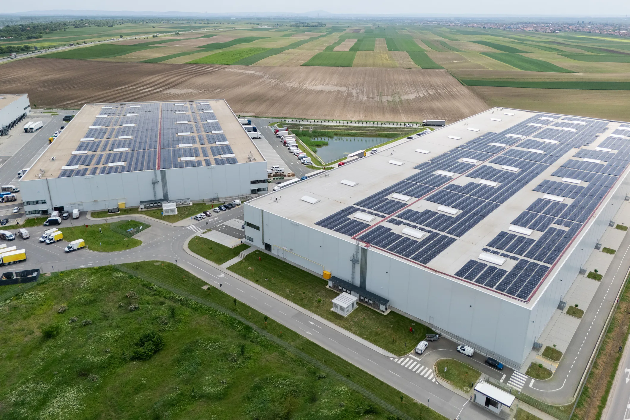 Aerial view of two large warehouses with solar panels on the roofs surrounded by roads and farmland.
