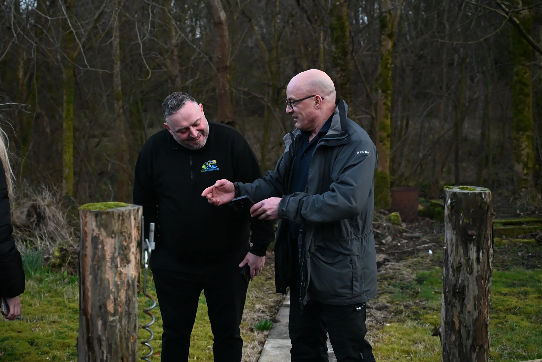 Two men outdoors in a wooded area, one showing something on a device to the other.