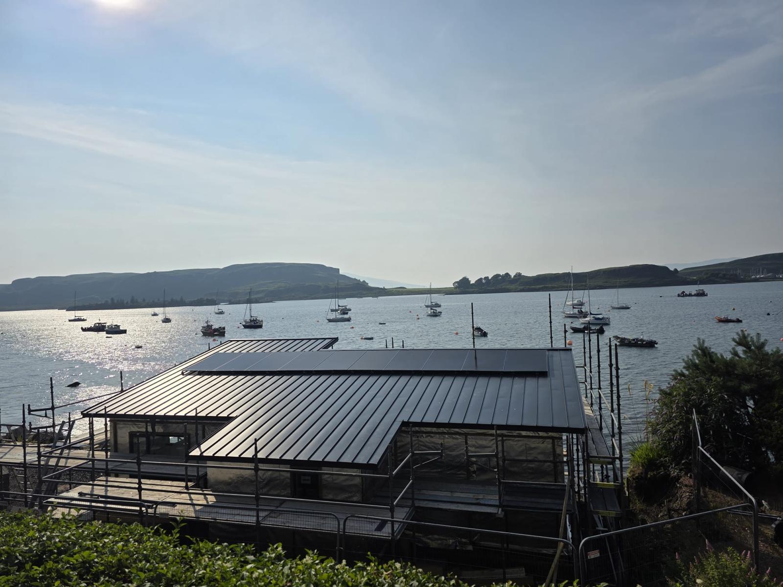 Metal roof building under construction by a calm body of water with boats and hills in the background.