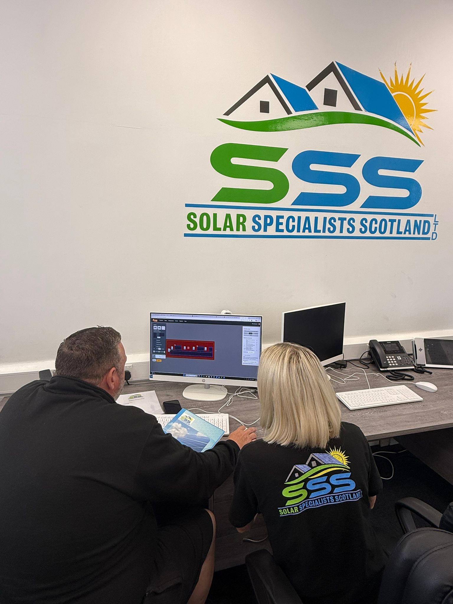 Two people sitting at a desk working on a computer under a Solar Specialists Scotland logo on the wall.