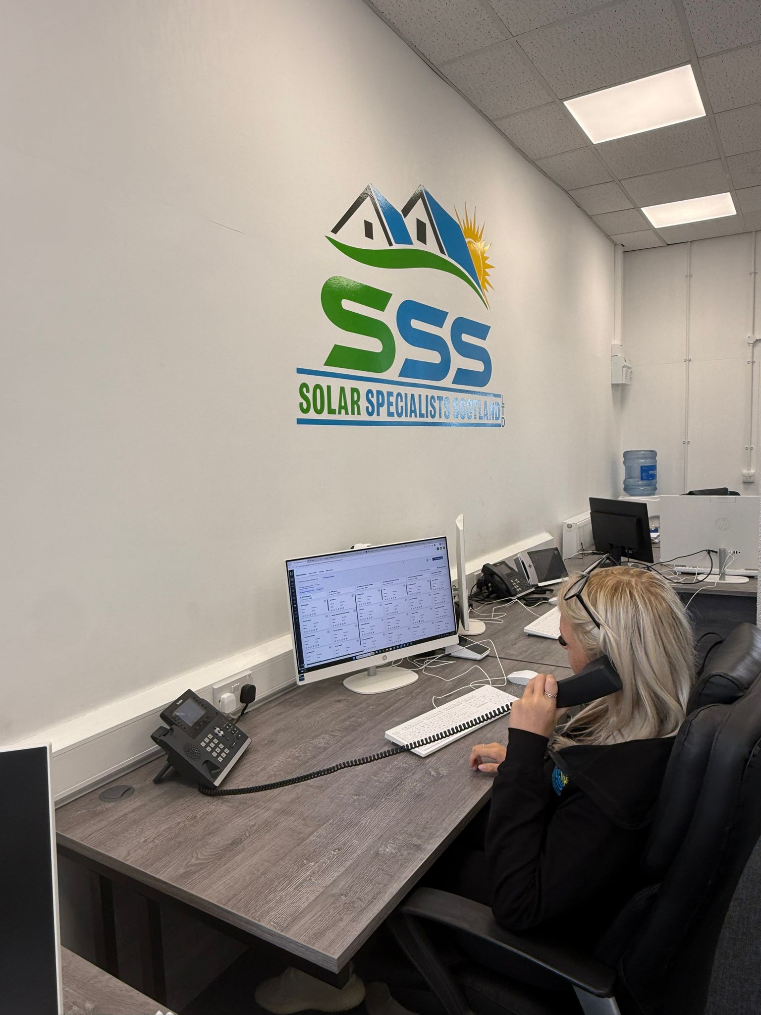 Woman with blonde hair on the phone sitting at a desk with a computer and Solar Specialists Scotland logo on wall.