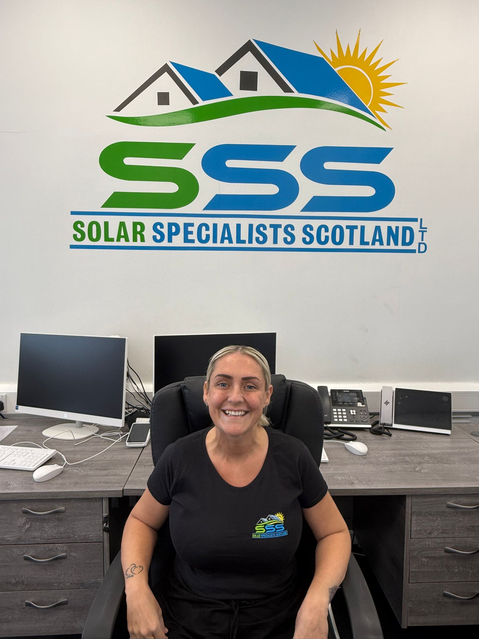 Smiling woman in black shirt with Solar Specialists Scotland logo seated in office chair before desk.