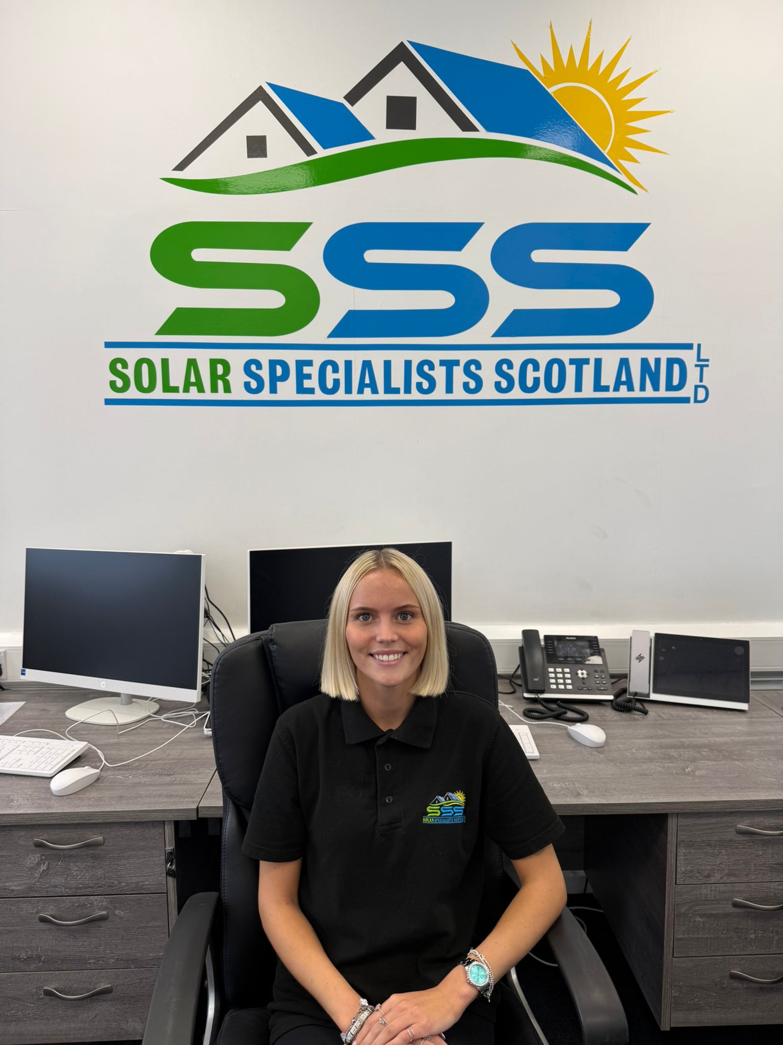 A smiling woman in black shirt with company logo sits at desk under Solar Specialists Scotland logo.