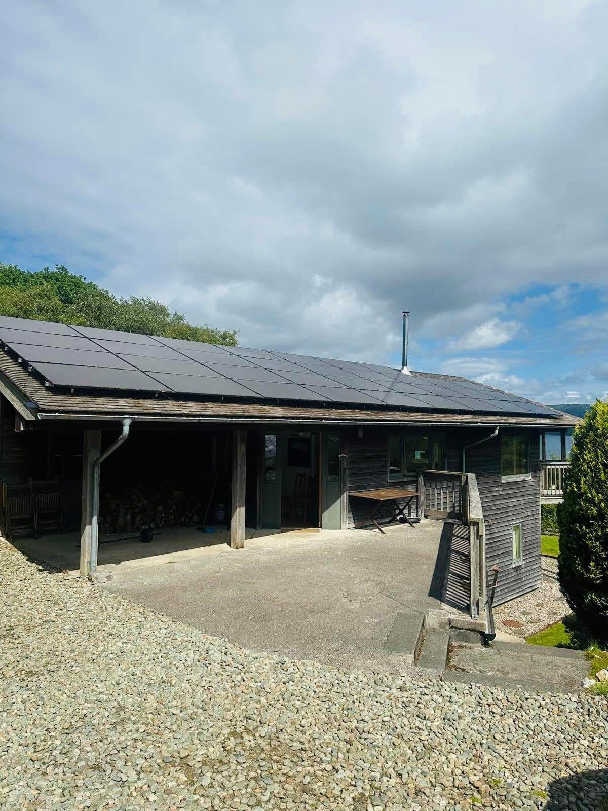 Wooden house with solar panels on the roof, concrete driveway, and gravel slope under a cloudy sky.