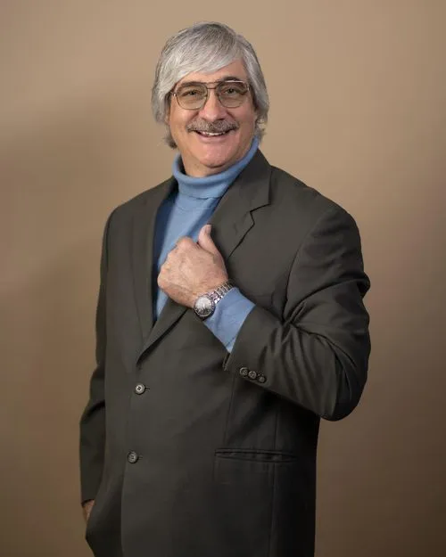 Smiling man with gray hair, wearing glasses, a blue turtleneck, and a dark blazer, posing against a beige background.