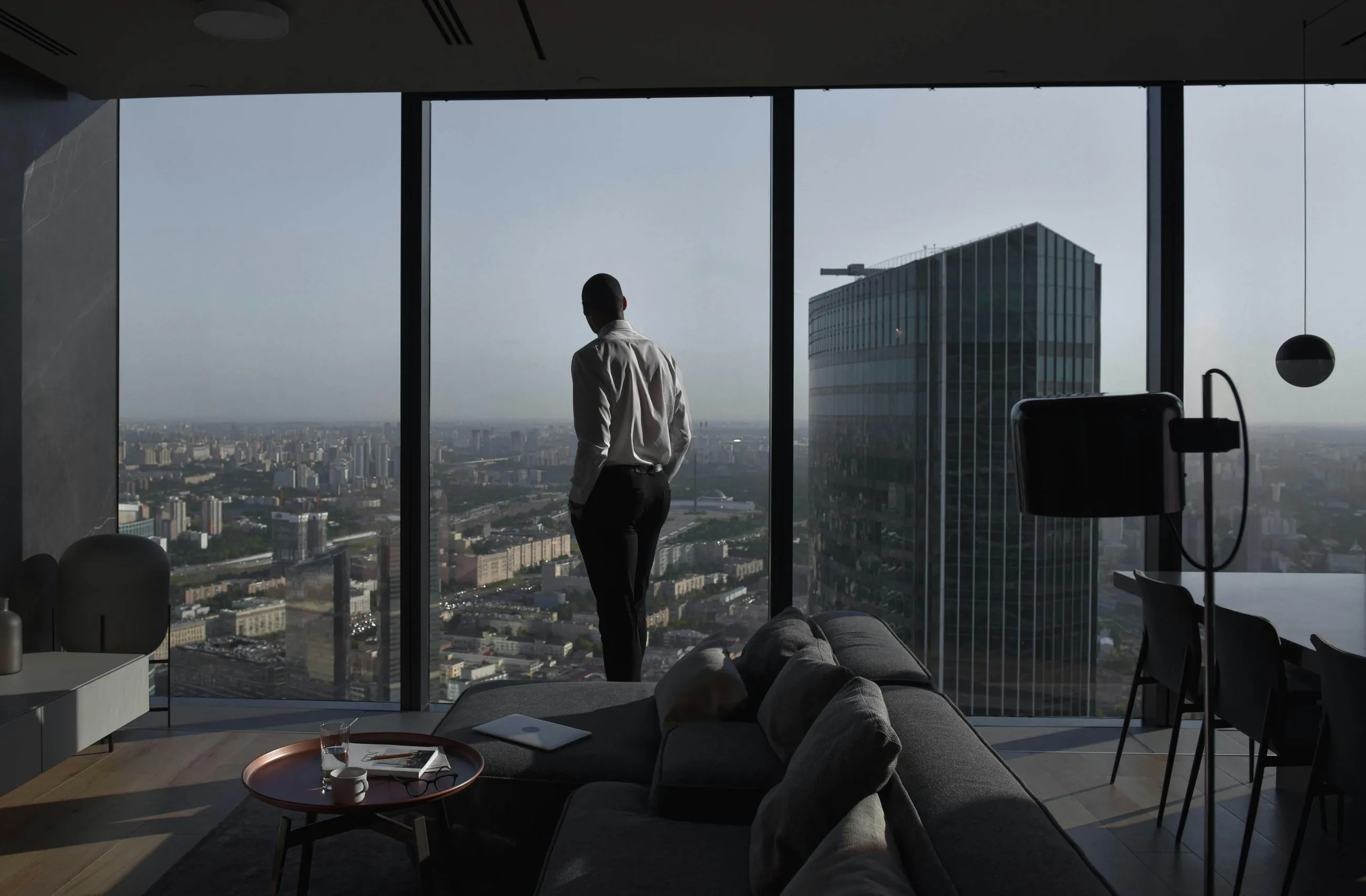 A man in a white shirt and black pants stands with hands in pockets looking out of a floor-to-ceiling window at a city skyline with a tall modern building nearby, inside a stylish living room.