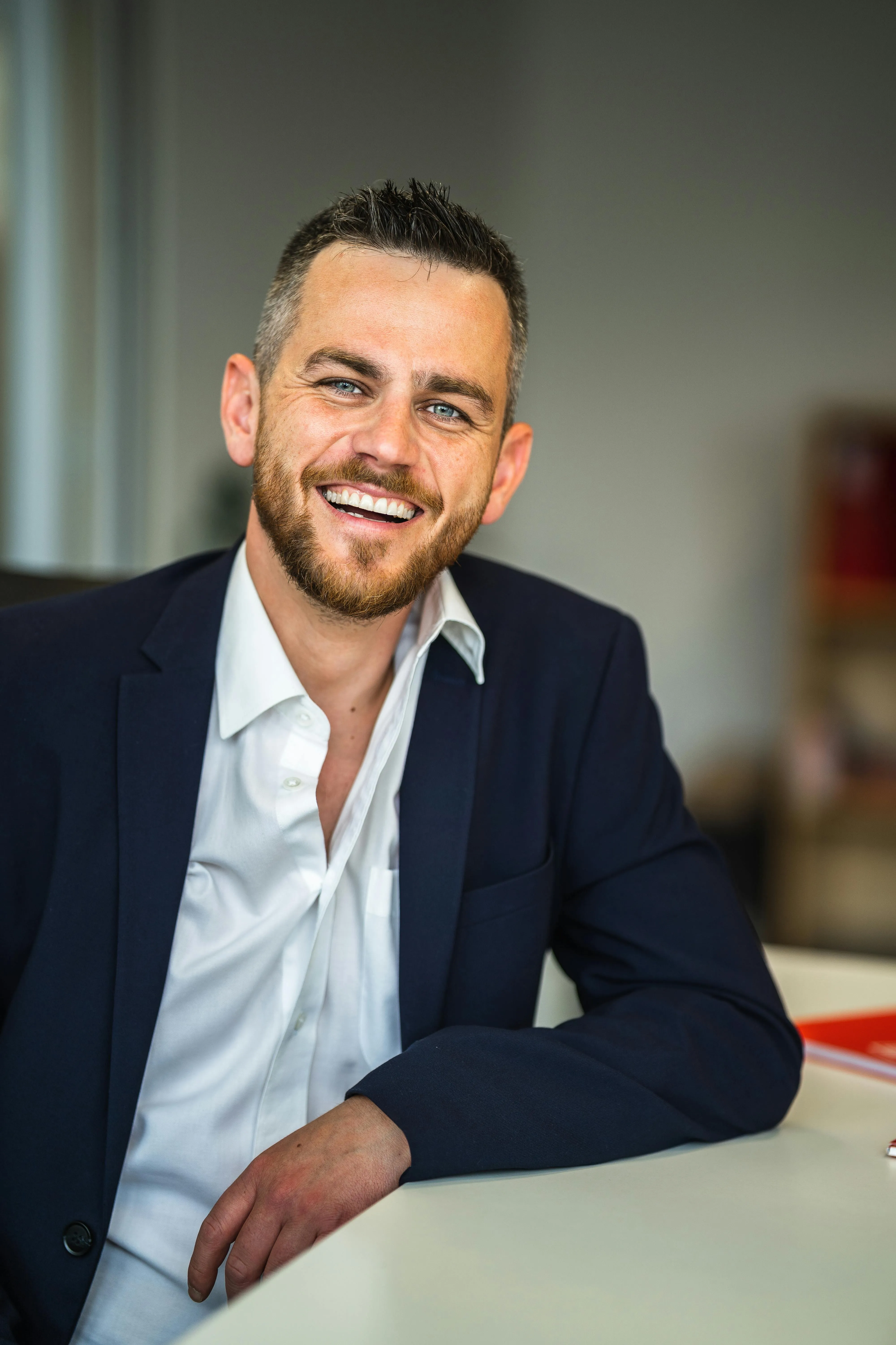 Smiling man with short hair and beard wearing a navy suit jacket and white shirt sitting at a desk.