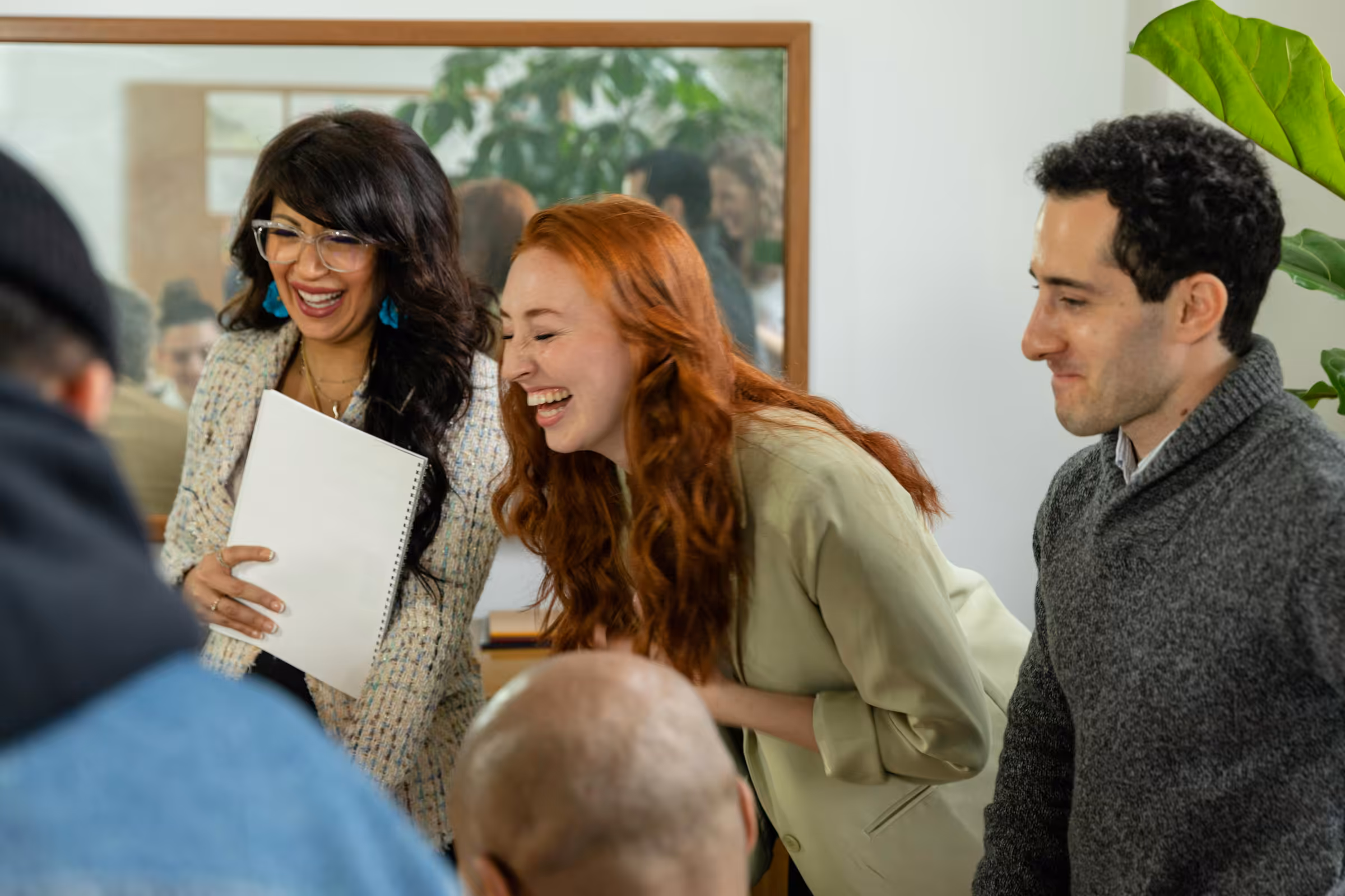 Three smiling adults engaged in conversation in a warmly lit room with plants and a mirror in the background.