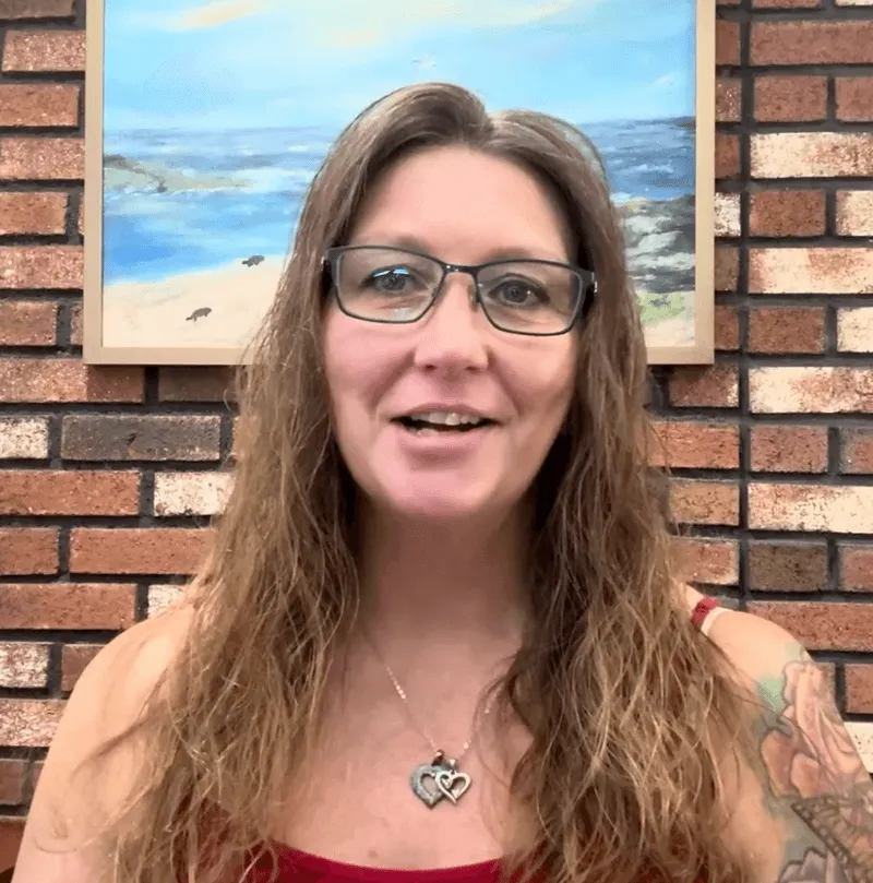 Woman with long wavy hair and glasses, wearing a red top and heart-shaped pendant, smiling indoors with a brick wall and a seaside painting background.