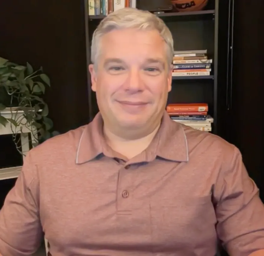 Smiling middle-aged man with short gray hair wearing a mauve polo shirt sitting in front of a bookshelf and a green plant.