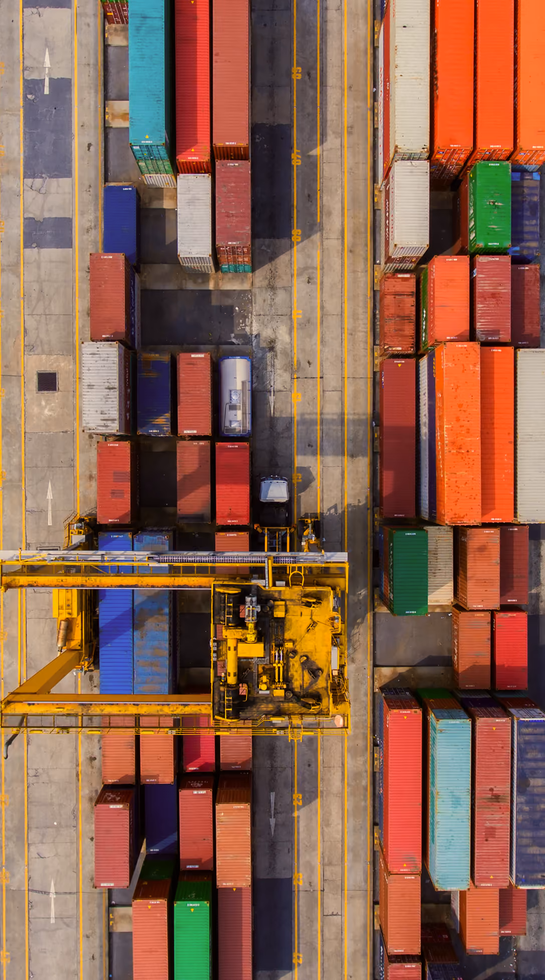 Aerial view of stacked colorful shipping containers and a yellow crane in a shipping yard.
