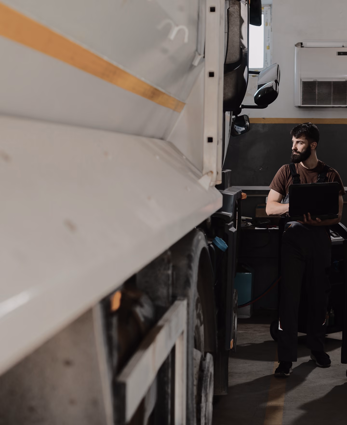 Bearded mechanic in overalls holding a laptop beside a large white truck in a garage.