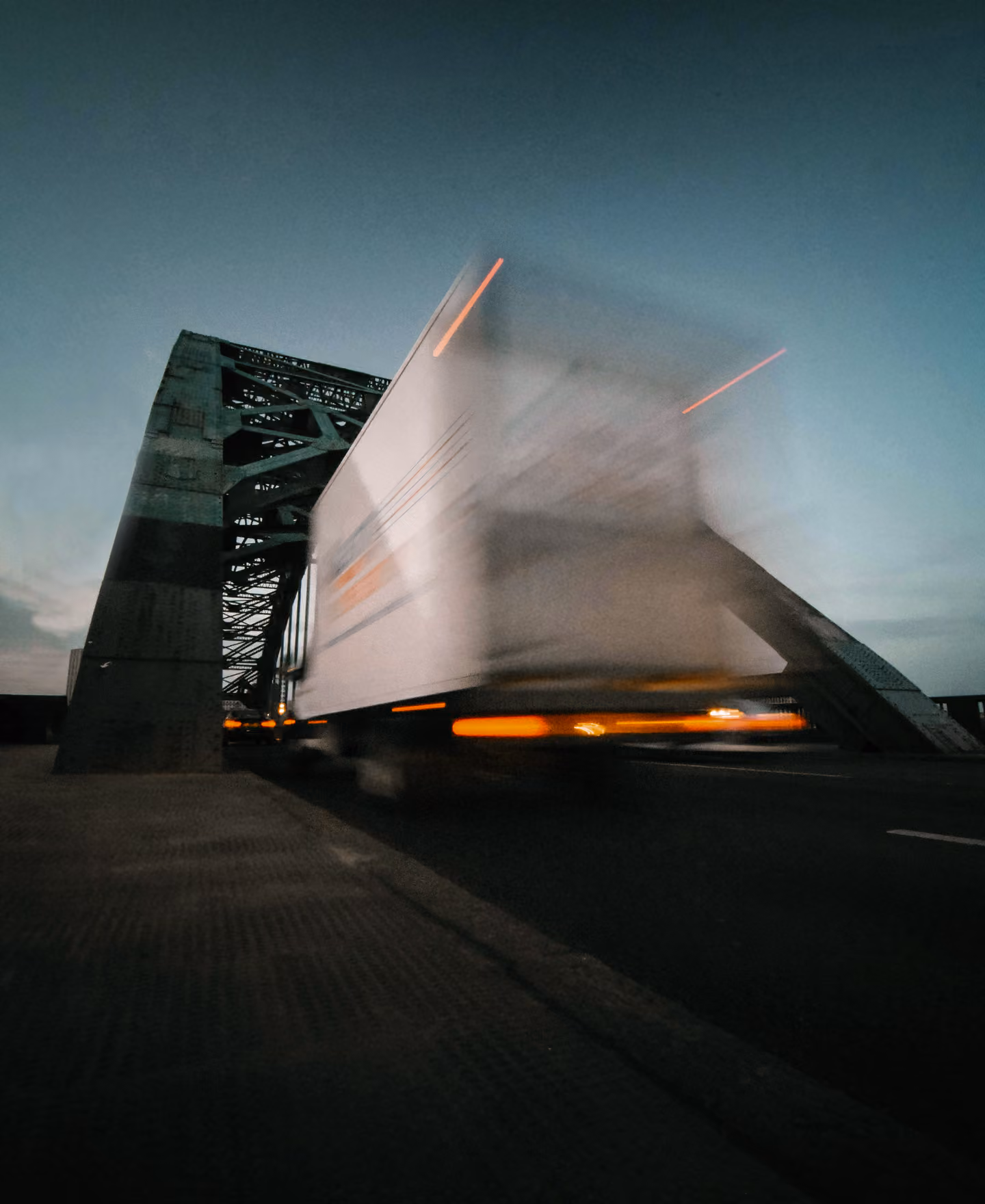 Blurred white truck speeding across a steel bridge at dusk with orange rear lights visible.