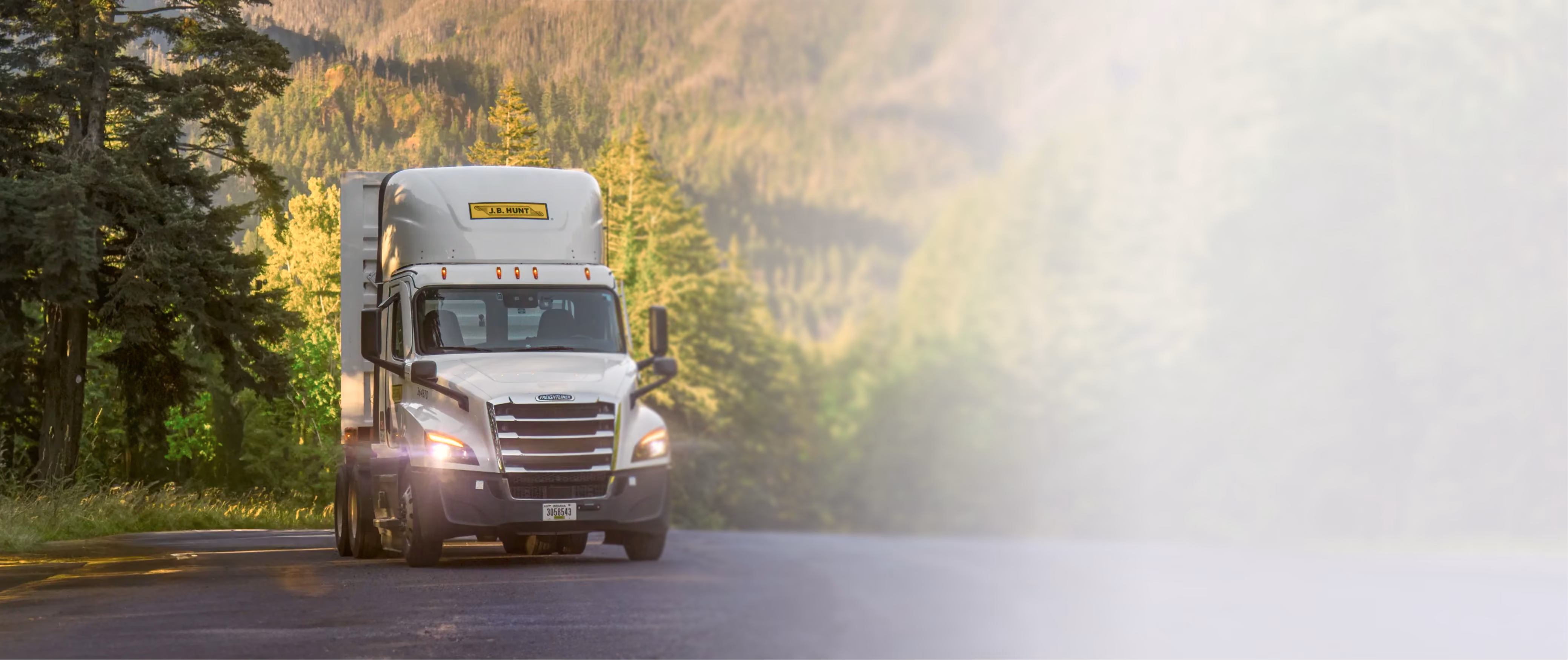 White Freightliner semi-truck driving on a forest road with mountains in the background.