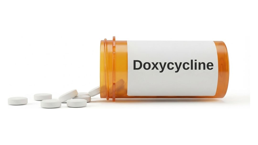An amber prescription bottle lying on its side on a white background with a plain white label reading Doxycycline, and six small round white tablets spilled out in front of it.