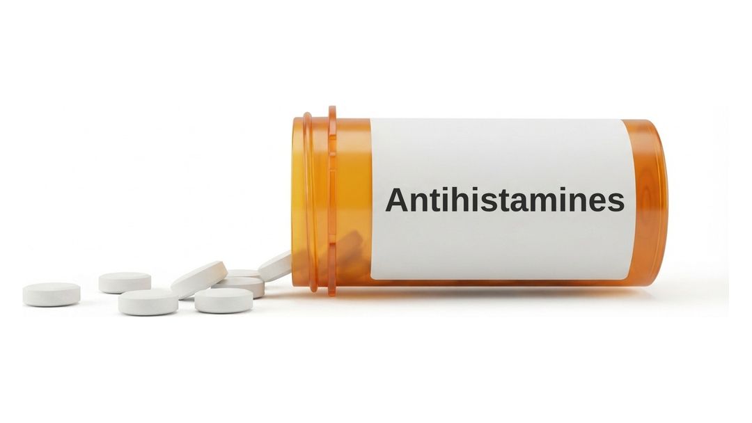 An amber prescription bottle lying on its side on a white background with a plain white label reading Antihistamines, and six small round white tablets spilled out in front of it.