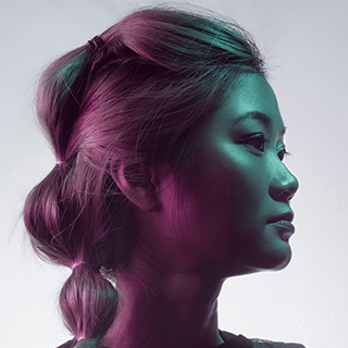 Side profile of a woman with styled hair lit by teal and pink lighting against a light background.