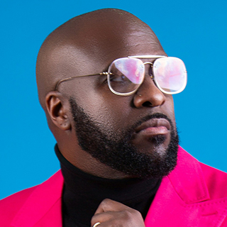 Man with beard wearing reflective sunglasses, black turtleneck, and bright pink blazer on blue background.