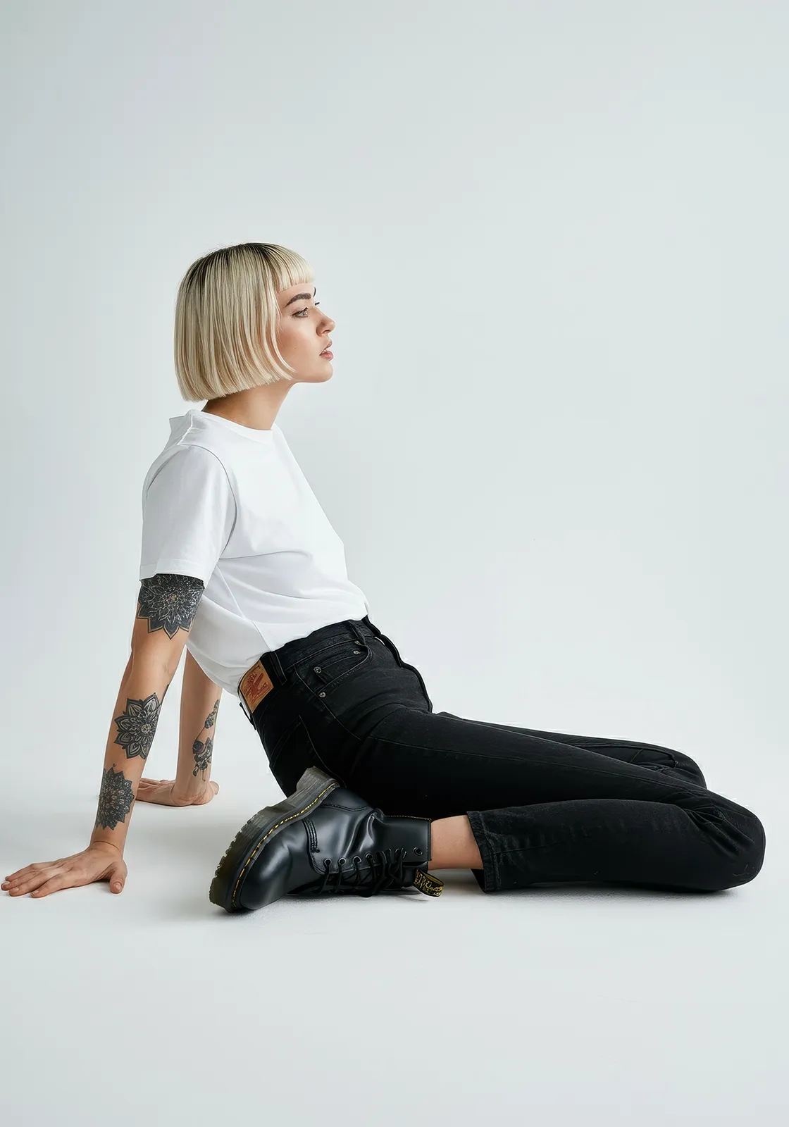 Woman with short blonde hair, tattoos, black jeans, and boots sitting on the floor, looking sideways.