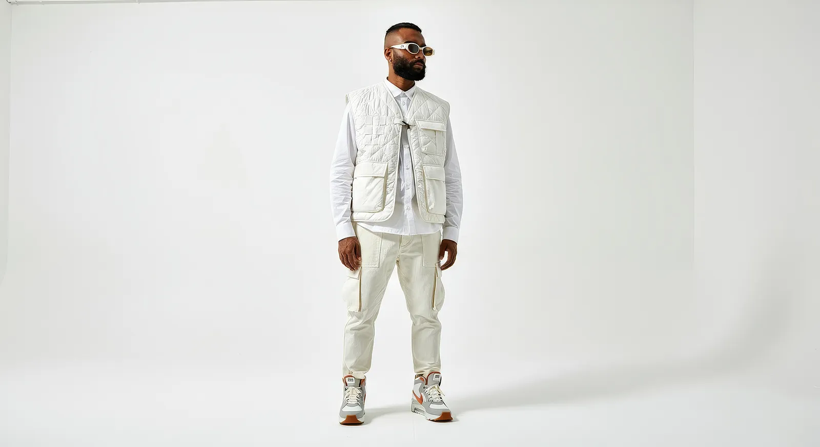 Man wearing all-white outfit with quilted vest, cargo pants, and sneakers, standing against a white backdrop.