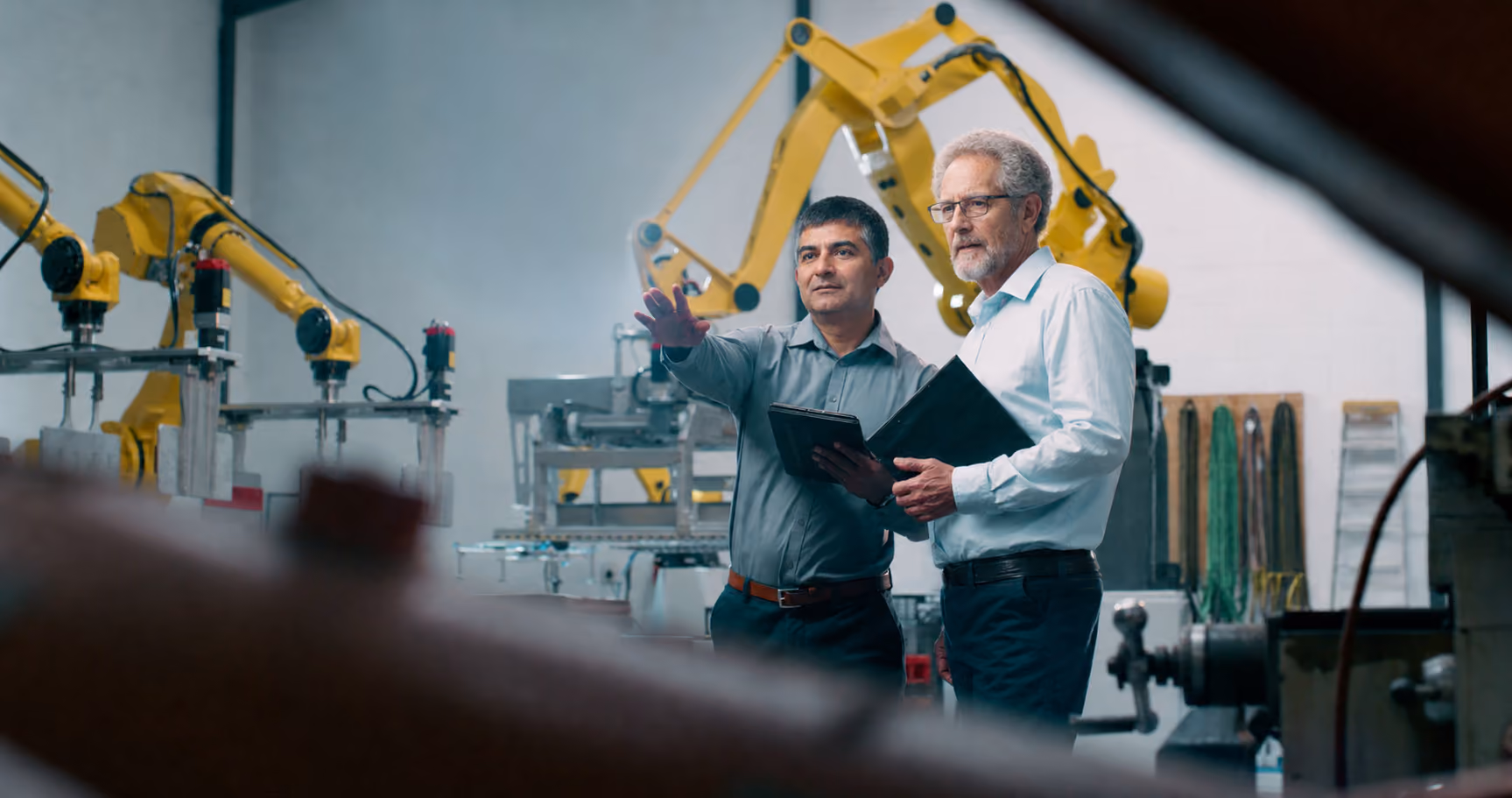 Two engineers discuss and point while holding a tablet in a factory with yellow robotic assembly arms.