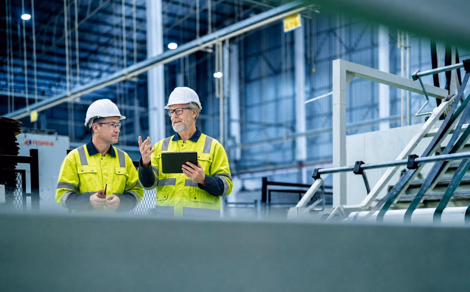 Two factory workers in yellow safety jackets and helmets discuss using a tablet inside a factory.