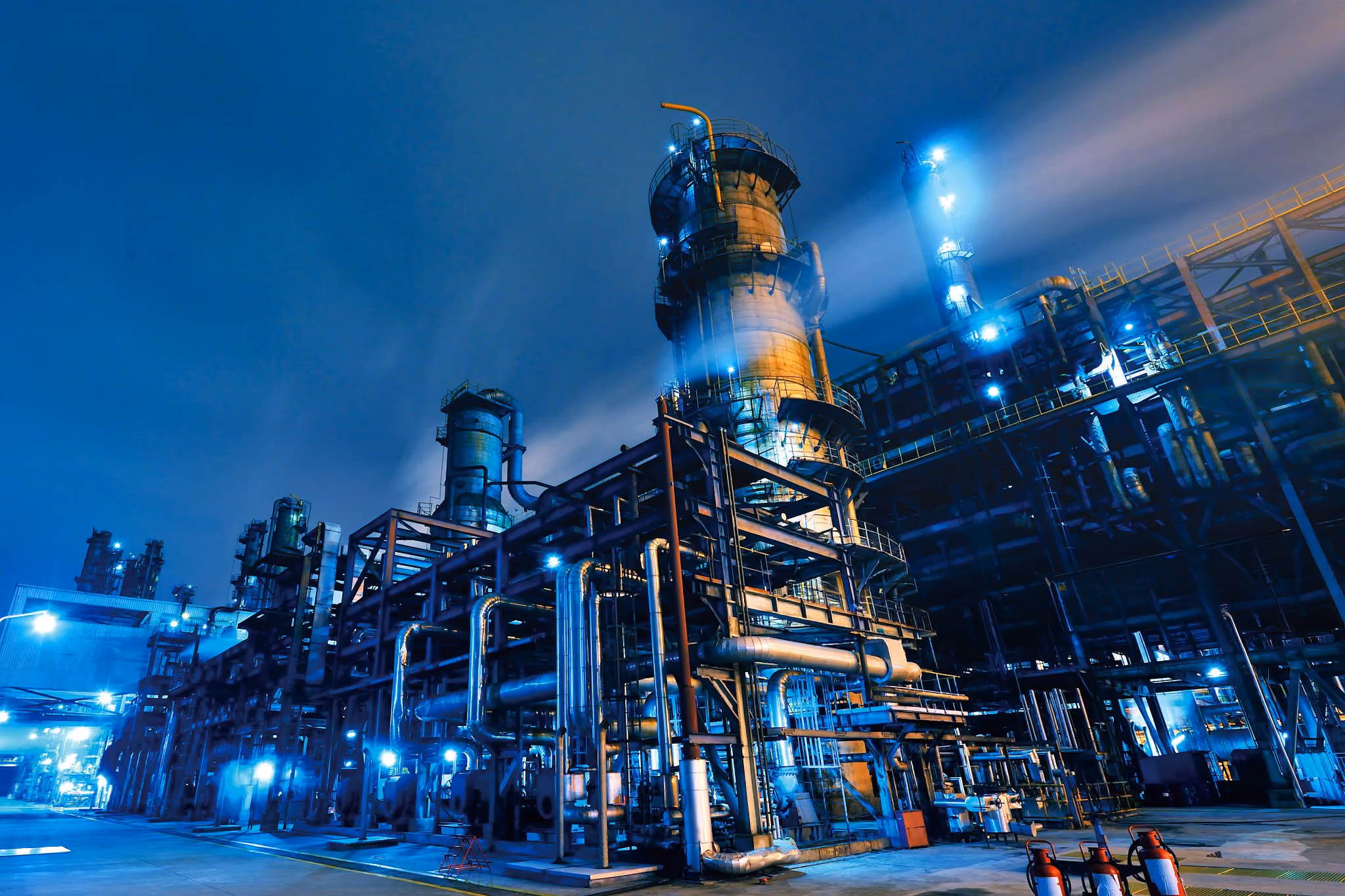 Industrial refinery plant at night illuminated with blue lights and visible piping and towers.