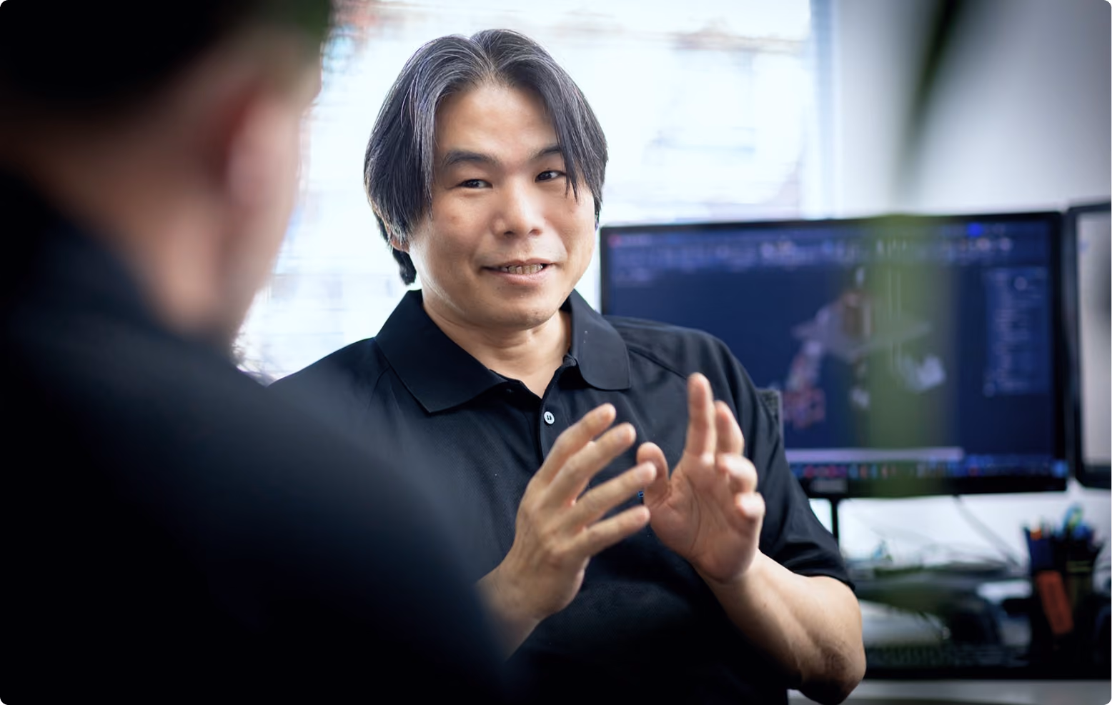 Man in a black shirt gesturing with hands while talking, with a computer screen in the background.