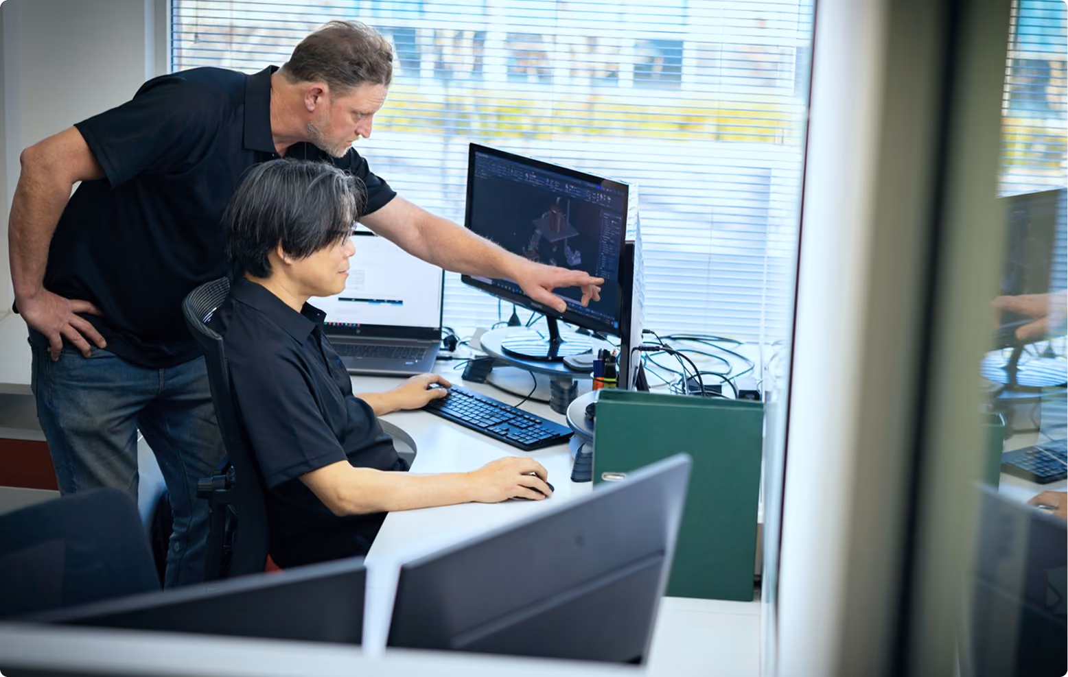 Man standing points at computer screen while seated man works at desk with two monitors.