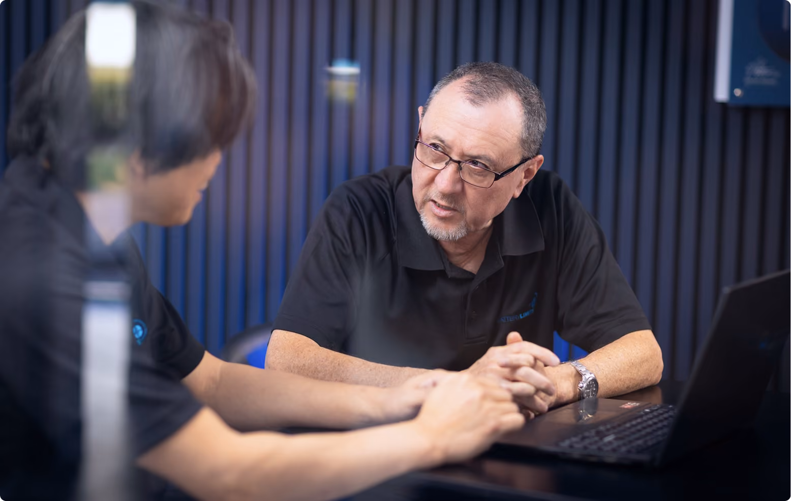 Two men having a discussion at a table with a laptop, one listening intently through glasses.