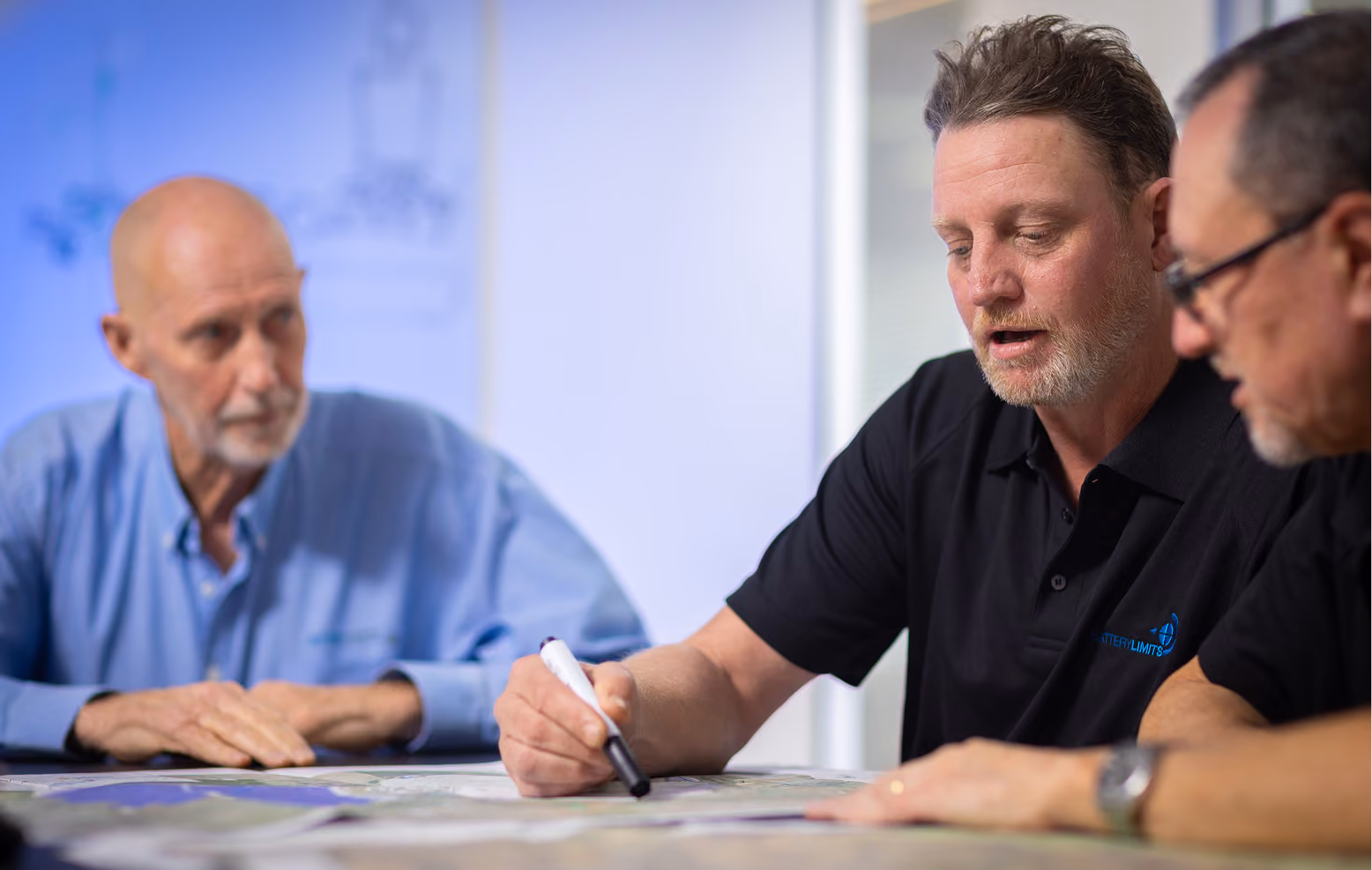 Three men around a table discussing documents; one man holds a marker and explains.