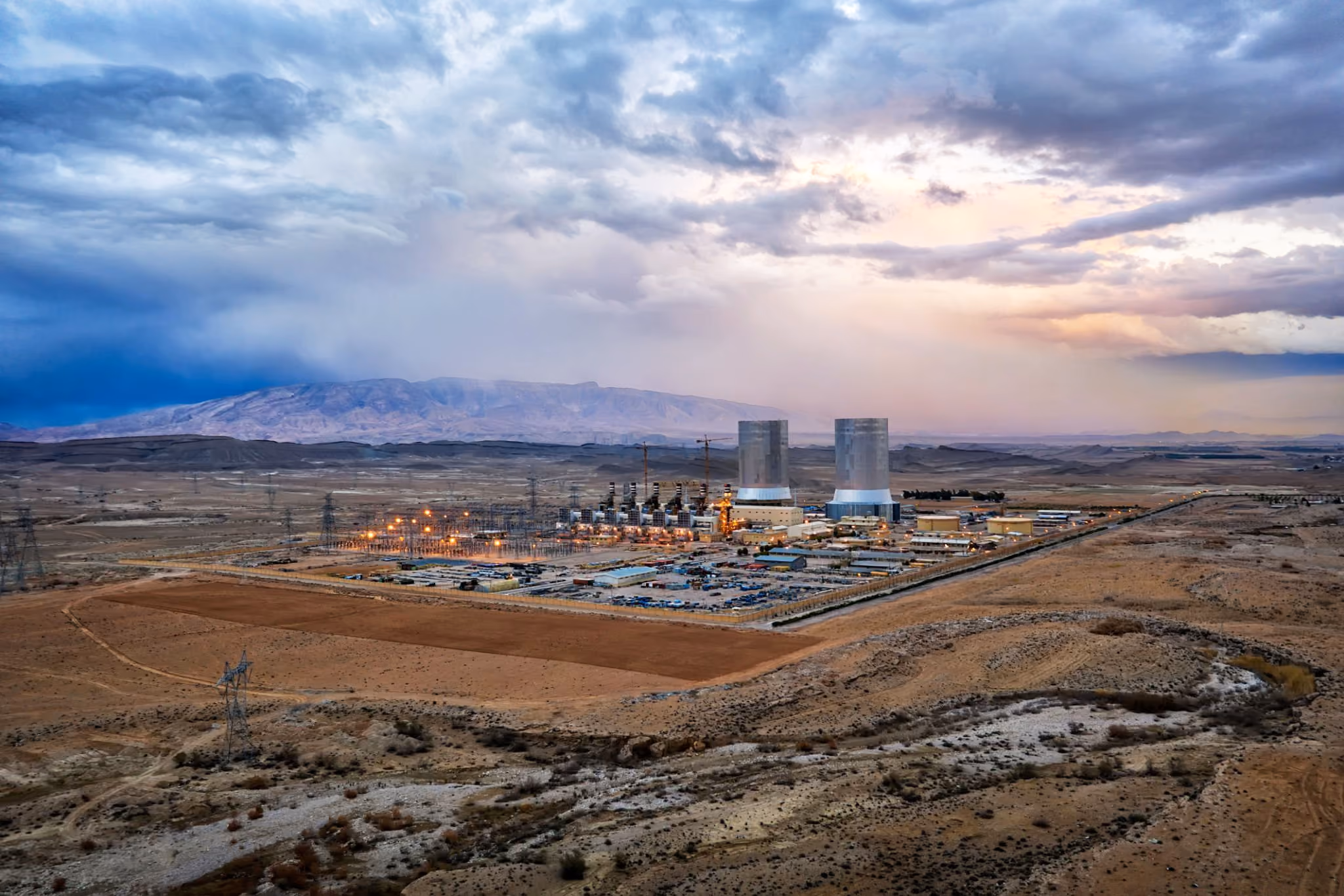 Power plant with two cooling towers in a desert landscape under a cloudy sky at dusk.