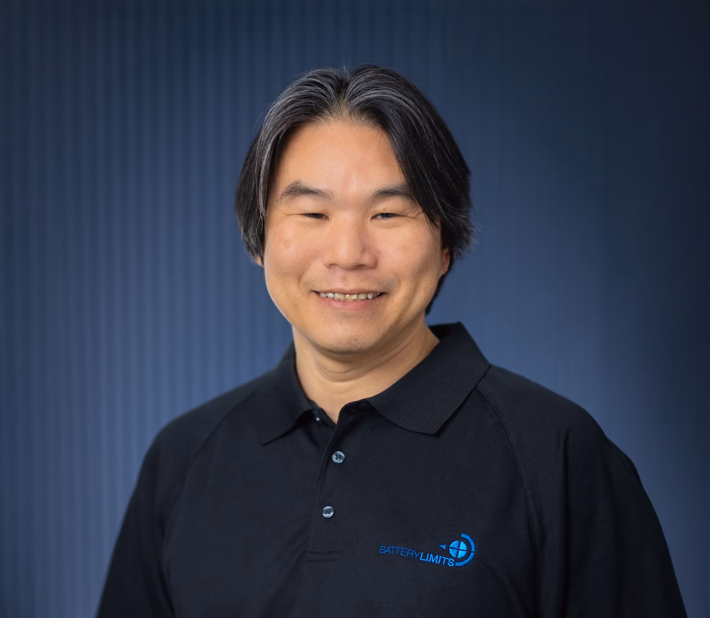Smiling man with black and gray hair wearing a black Battery Limits polo shirt against a dark blue background.