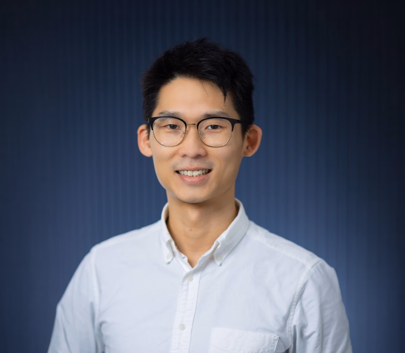 Smiling young man wearing glasses and a white buttoned shirt against a dark blue background.