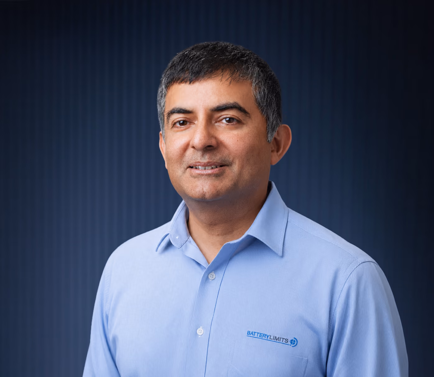 Man with short dark hair wearing a light blue shirt with Battery Limits logo on a dark background.