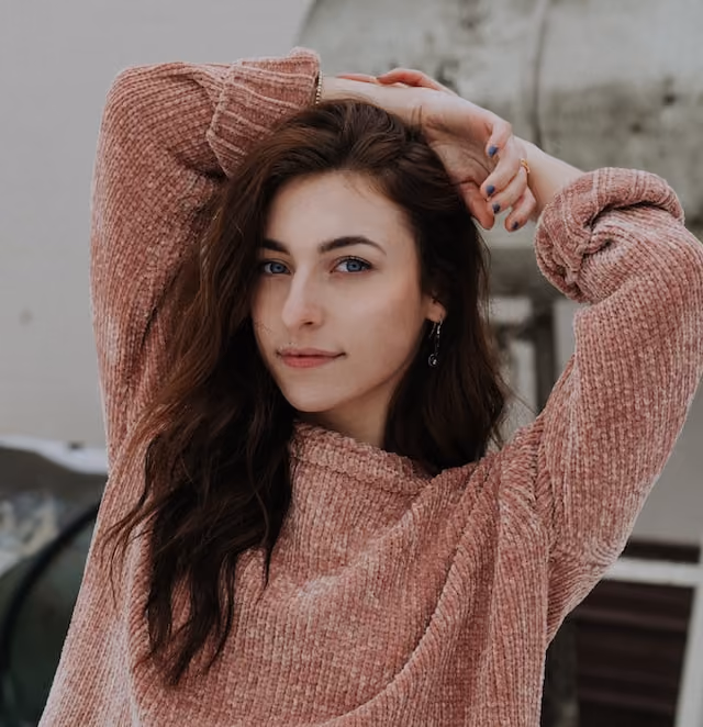 Young woman with long dark hair in a pink sweater posing with hands above her head Image.