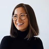 Smiling woman with glasses and shoulder-length dark hair wearing a black turtleneck against a white background.