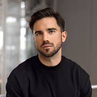 Man with short dark hair and beard wearing a black shirt in front of a blurred background.