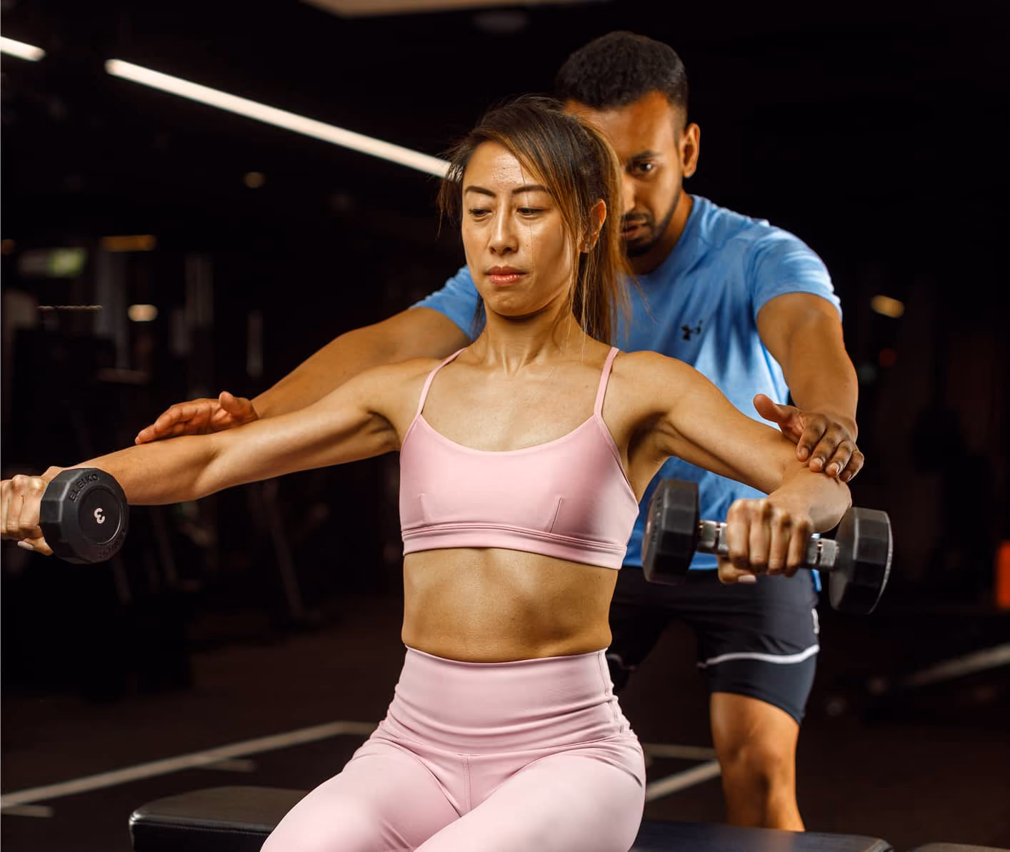 Woman lifting dumbbells for shoulder exercise with male trainer guiding her arm movements.