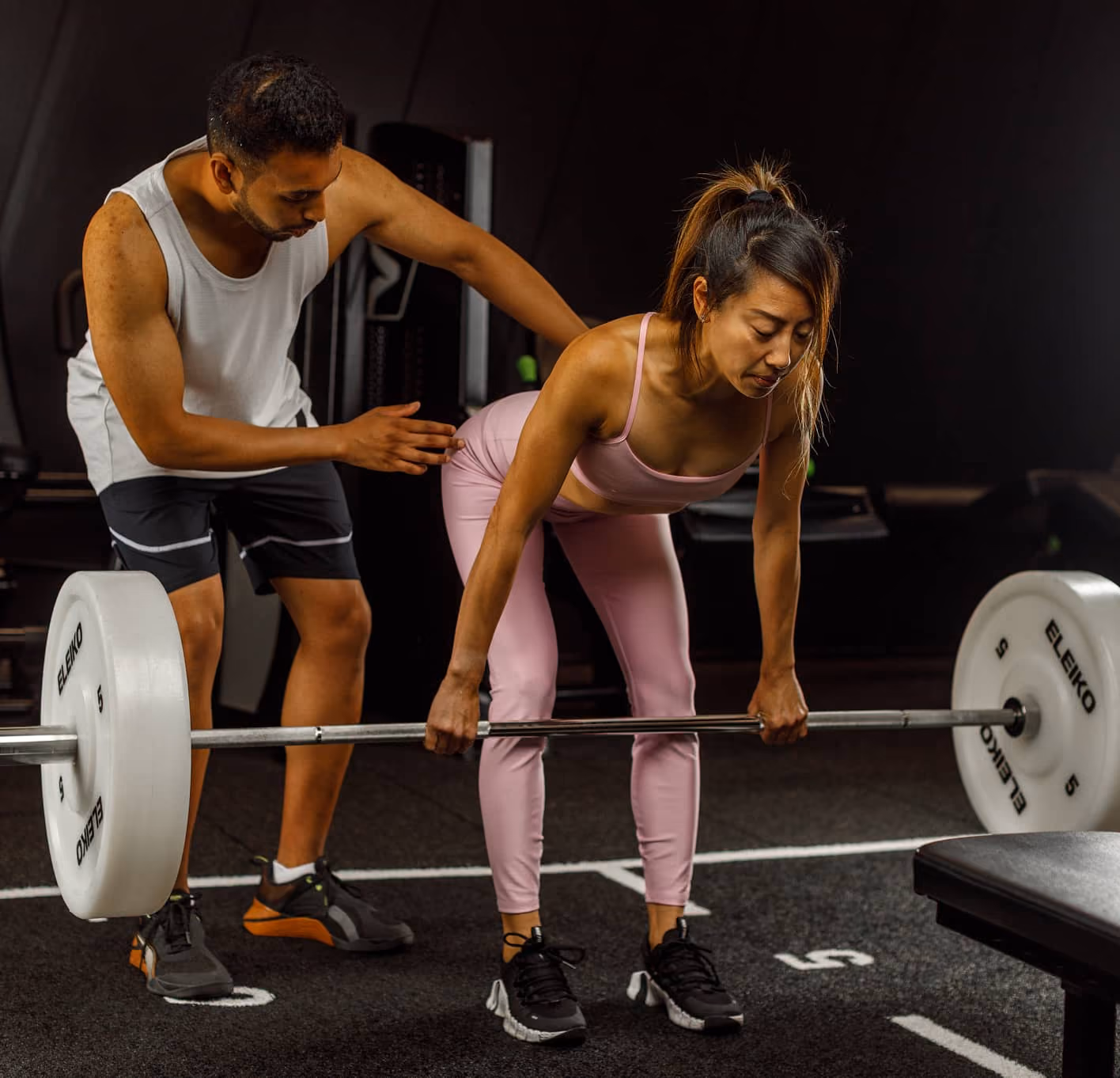 Trainer guiding a woman lifting a barbell with weights in a gym for proper form.