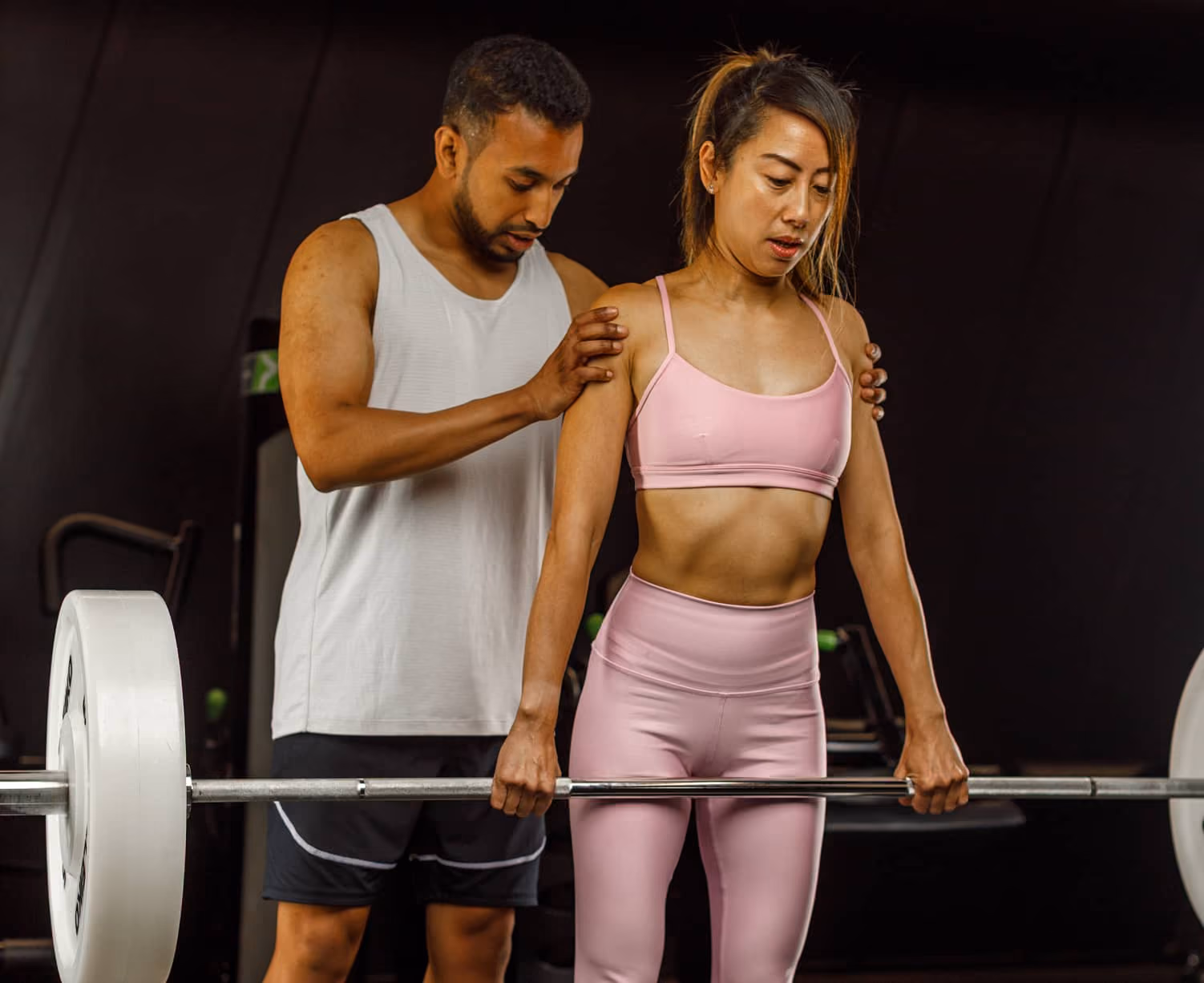 Man in white tank top coaching woman in pink workout clothes lifting a barbell at gym.