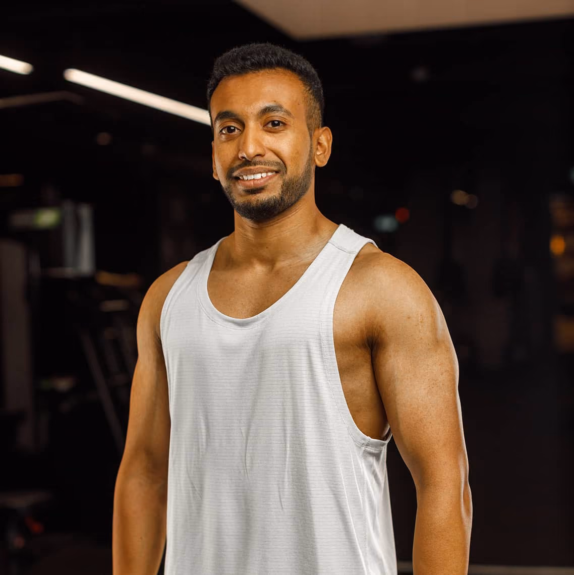 Smiling muscular man in a white tank top standing in a dimly lit gym.